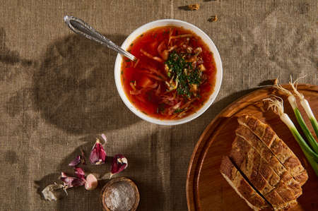 White ceramic bowl with borscht, green onions and sliced bread on cutting wooden board, peeled garlic and container of salt on linen tablecloth. Flat lay food background. Ukrainian traditional cuisineの写真素材