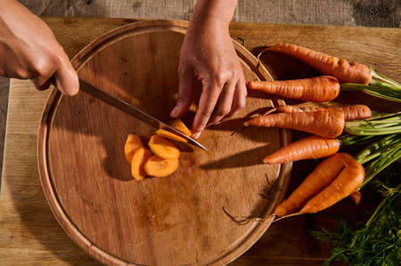 Top view of female hands cutting, chopping, slicing fresh raw baby carrot on a wooden board. Close-up of hand with knife cutting fresh vegetable. Flat lay food backgroundの写真素材