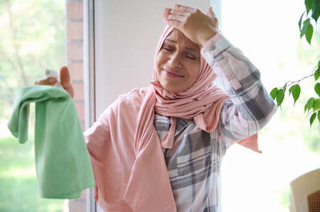 Sorrowful Arab Muslim woman in hijab, housewife holds her head from fatigue, closes her eyes from frustration and many household chores, holding a cleaning spray and a rag while cleaning windowsの写真素材