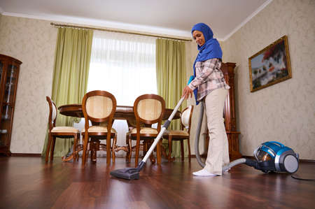 Beautiful Arab Muslim woman with head covered in hijab cleaning floor with a vacuum cleaner in the luxurious living room. Concept of easy cleaning, cleanup, household, housekeepingの写真素材