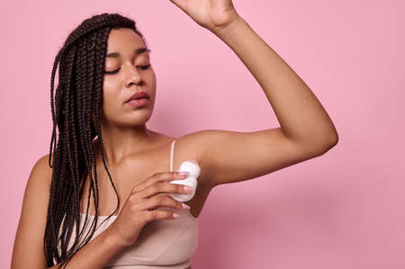 Close-up isolated beauty portrait on pink background of African young woman with dreadlocks in beige underwear applying antiperspirant under the armpits. Purity, hygiene, body care concept. Copy spaceの写真素材