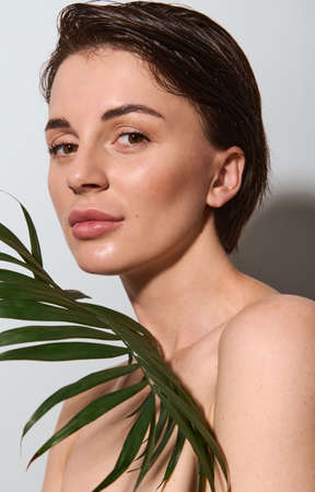 Gorgeous delightful beautiful young brunette of natural beauty poses nude with exotic palm leaf on a white background. Skin and body care, wellness and well-being conceptの写真素材