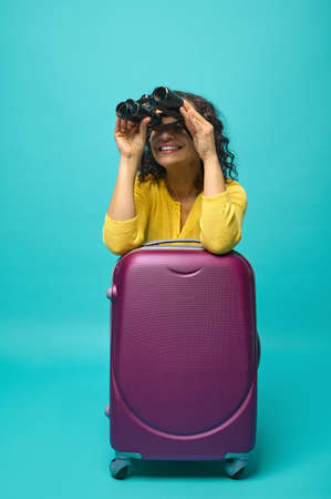 Young woman traveler adventurer explorer tourist in bright yellow clothes leaning on a suitcase and smiles toothy smile looking through binoculars, isolated on blue background with copy spaceの写真素材
