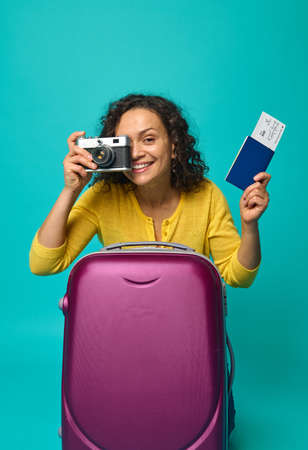 Cheerful smiling beautiful woman holding passport with ticket and boarding pass and a vintage camera, photographing, smiling toothy smile looking at camera against blue colored background. Copy spaceの写真素材
