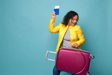 Funny joyful woman traveler tourist with curly hair, dressed in yellow jacket raised her arms with passport and ticket and dances with a suitcase in her hands, enjoying her vacation. Travel conceptの写真素材