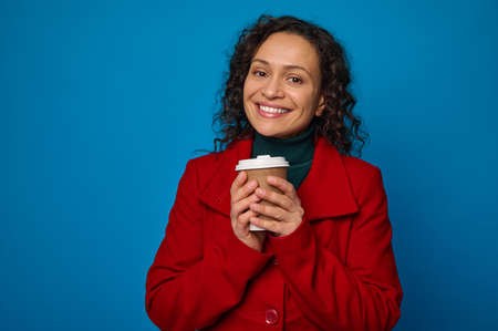 Charming dark-haired woman in bright colorful clothes warming her hands, holding a takeaway paper...