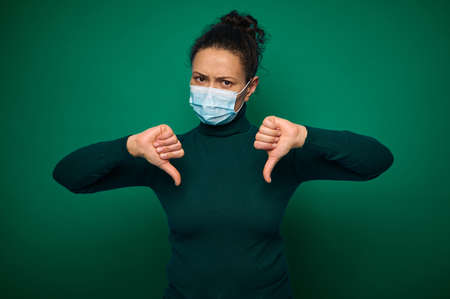 Isolated portrait on green background of a serious millennial woman in protective medical mask,...