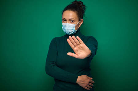 Isolated portrait on green background of a serious millennial woman in protective medical mask,...