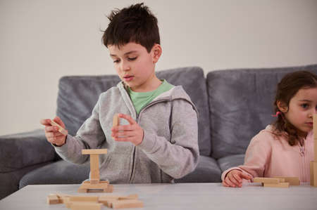 Creative children, boy and cute girl focused on building from wooden blocks, playing developmental board games, building complex tall structures at home. Educational leisure and family pastime conceptの写真素材