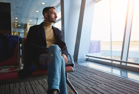 Handsome middle aged confident businessman on business trip, sitting in the departure lounge, admiring the view from the panoramic windows of the international airport to the runway with airplanesの写真素材