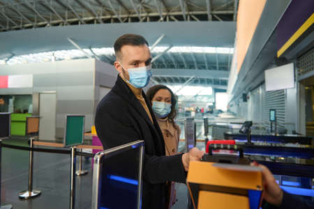Young couple of business partners in protective medical masks, traveling during pandemic, standing at the check-in counter, passing customs and passport control at the airport terminal. Copy ad spaceの写真素材