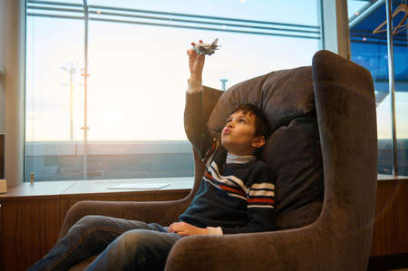 Handsome European teenage boy plays with toy airplane sitting in a chair by panoramic windows overlooking the runway at sunset while waiting to board flight at international airport departure terminalの写真素材
