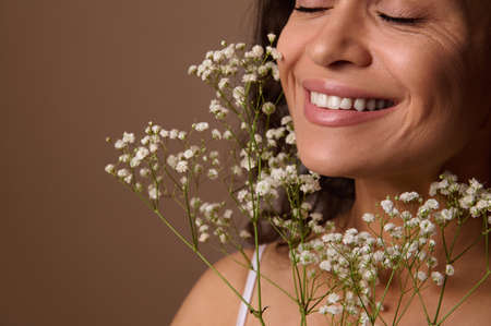 Close-up portrait of Hispanic pretty woman with closed eyes and beautiful smile, holding Gypsophila white sprig against beige background. Femininity, sensuality, natural beauty, Women's Day conceptの写真素材