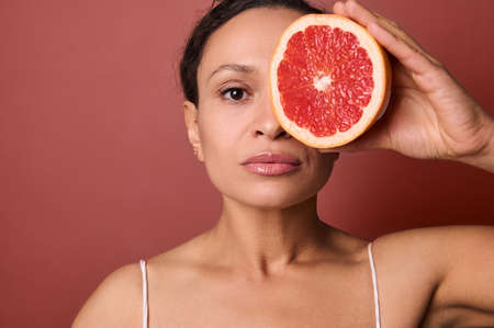 Attractive woman with perfect healthy skin holds a grapefruit in front of her eye, covering with it...