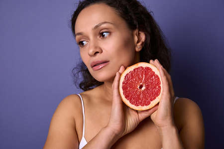 Close-up. Beautiful dark-haired woman with a half of red fresh juicy grapefruit near her face, looking away isolated over purple background with copy ad space. Health, body and skin care conceptの写真素材