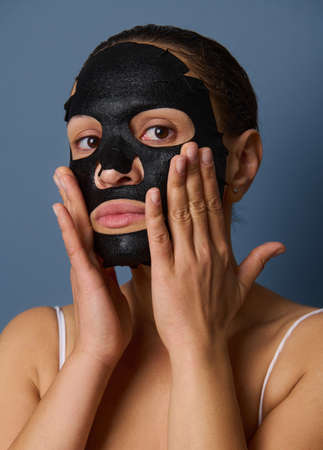 Close-up portrait of beautiful woman looking at the camera while putting on a nourishing smoothing moisturizing black face mask taking care of the beauty of her skin. Isolated on blue-gray backgroundの写真素材