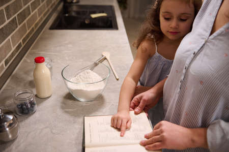 Beautiful happy child, adorable little girl points with her finger on a recipe book, standing at kitchen countertop close to her mom while enjoying cooking together, preparing dough for Shrove Tuesdayの写真素材