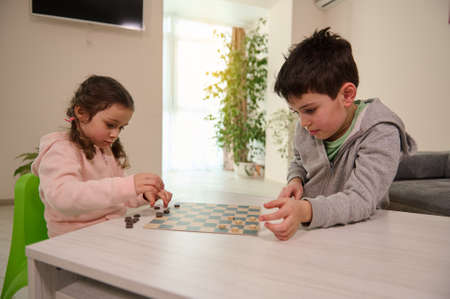 Two adorable Caucasian elementary aged kids, boy and girl, brother and sister having great time playing checkers board game together at home interior.の写真素材