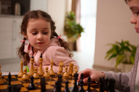 Close-up portrait of a beautiful cute little European girl playing chess game with her elder brother who picks up a chess piece and make his move. Intellectual smart development board games for kidsの写真素材