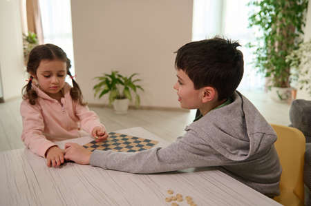 Two adorable Caucasian elementary aged kids, boy and girl, brother and sister having great time playing checkers board game together at home interiorの写真素材
