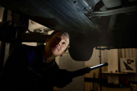 Technician checking modern car at automobile repair shop. Auto mechanic using a flashlight lamp for...