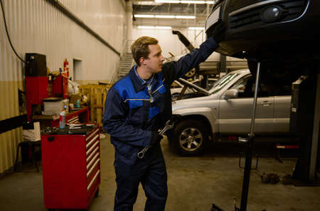 Handsome young mechanic in professional uniform, holding a wrench and working in auto service with...