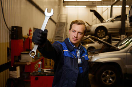 Professional portrait of a young Caucasian auto mechanic in uniform holding a wrench while standing...