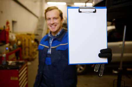 Focus on white blank sheet with copy ad space on a clipboard in the hand of a blurred smiling technician, car engineer, mechanic standing in the garage of repair shop. Car maintenance conceptの写真素材
