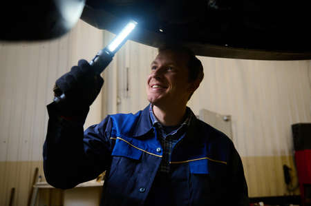 Mechanic, technician, auto engineer checks the underbody of a lifted car with flashlight in the garage of a repair shop. Car warranty repair and maintenance, car service conceptの写真素材