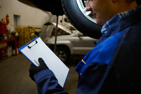 Rear view of auto mechanic, technician doing checklist on clipboard with white blank paper sheet, while inspecting the car lifted in the lift in repair shop. Car maintenance conceptの写真素材