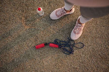 Overhead view of female athlete legs wearing pink sneakers and standing on an asphalt treadmill next to a lying down skipping rope and water bottleの写真素材