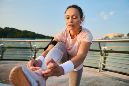 Female athlete with earphones, runner tying sneakers laces while enjoying morning jog on a city bridge treadmill. Run, jogging, healthy active lifestyle, cardio workout and outdoor fitness conceptの写真素材