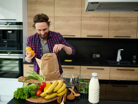 Handsome happy Caucasian man unpacking purchased healthy food standing by a kitchen countertop with bottle of milk and healthy raw vegan vegetables and fruits on a wooden cutting boardの写真素材