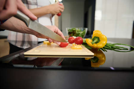 Close-up of man's hands slicing tomatoes and yellow bell pepper on a cutting board on the background of a pregnant woman holding sprig with fresh tomatoes in the home kitchen. Healthy eating conceptの写真素材