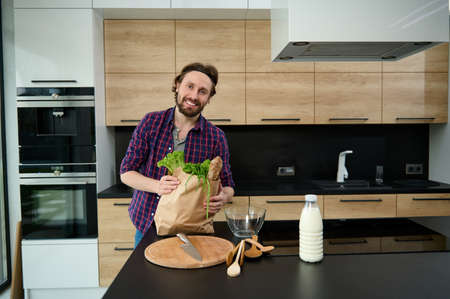 Portrait of a handsome charming European guy with eco papr bag full of healthy food, greens and whole grain bread standing by a kitchen table in his spacious smart villaの写真素材