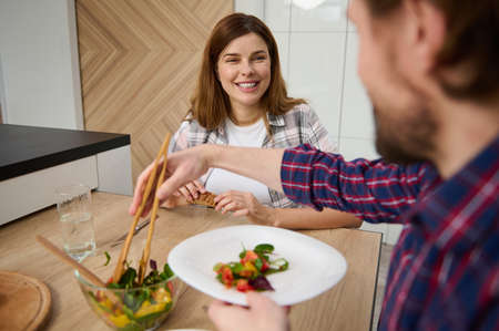 Beautiful happy young Caucasian couple in love, enjoying a dinner together, having fun discussion and laughing while sitting at table in the house with beautiful light interiorの写真素材