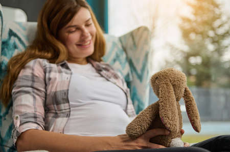 Delightful pregnant woman sits with a plush rabbit, dreams of future child at home near window overlooking garden. Cute woman in anticipation of baby enjoys her carefree pregnancy and maternity leaveの写真素材