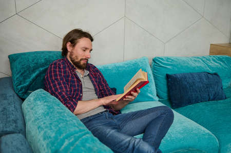 Serene confident concentrated young bearded Caucasian man relaxing on the couch, reading book in home environment. Education, study, knowledge and erudition conceptの写真素材