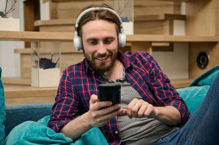 Close-up portrait of a happy Caucasian young man using social media at home. Handsome guy with wireless headphones relaxing on the couch at home. Hobby, entertainment, leisure activity conceptの写真素材