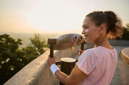 Sporty pretty woman looking through a telescope at the city. Travel and Tourism concept. Woman using...