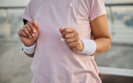 Cropped image of an unrecognizable female athlete in pink t-shirt with hands wearing white terry wristbands holding earphones while exercising outdoor early in the morning. Physical activity conceptの写真素材
