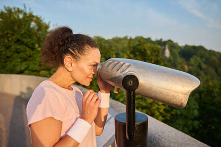 Middle aged woman looks through a telescope at the city. Athletic woman in a pink t-shirt is watching the beautiful green nature and the river background through stationary city binocularsの写真素材