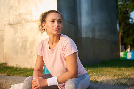 Confident portrait of an athletic woman, sportswoman in pink t-shirt and gray leggings sitting on green grass outdoors, looking away, relaxing after morning cardio workout outdoorsの写真素材