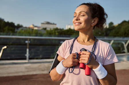 Happy confident sportswoman with a jumping rope, posing with closed eyes, standing on the modern glass city bridge, enjoying early sunny morning cardio workout outdoor. Cardio workout conceptの写真素材