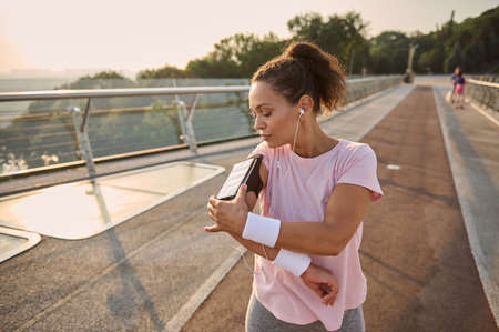 Sportswoman, African American female athlete runner adjusting mobile applications on her smartphone in a holder on her hand while exercising outdoor, standing on a treadmill on the city bridge.の写真素材