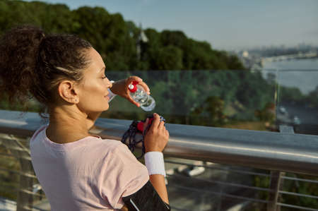 Rear view of a female athlete with a bottle of water in her hand after intense cardio workout and morning jog, looking away resting on a city bridge at dawn. Outdoor workout conceptの写真素材