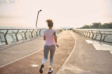 Rear view of young athletic woman, jogger, runner practicing sport outdoors, running fast along the...