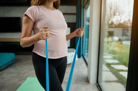 Cropped image of beautiful determined active pregnant woman, in anticipation of a baby, doing stretching exercises on arms with an elastic strap at home. Sport and pregnancy, healthy lifestyle conceptの写真素材