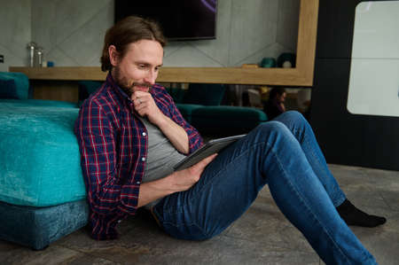 Relaxed bearded Caucasian man in casual denim and checkered shirt reads news on digital tablet,...