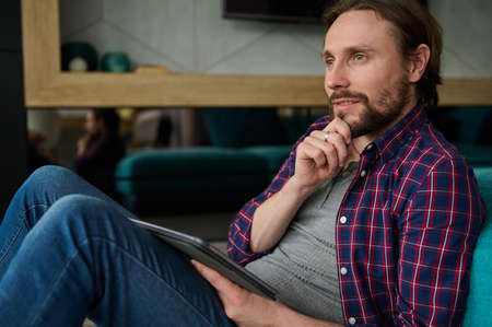 Pensive handsome bearded Caucasian man in casual denim and checkered shirt holding a digital tablet and looking thoughtfully away, sitting at home environmentの写真素材